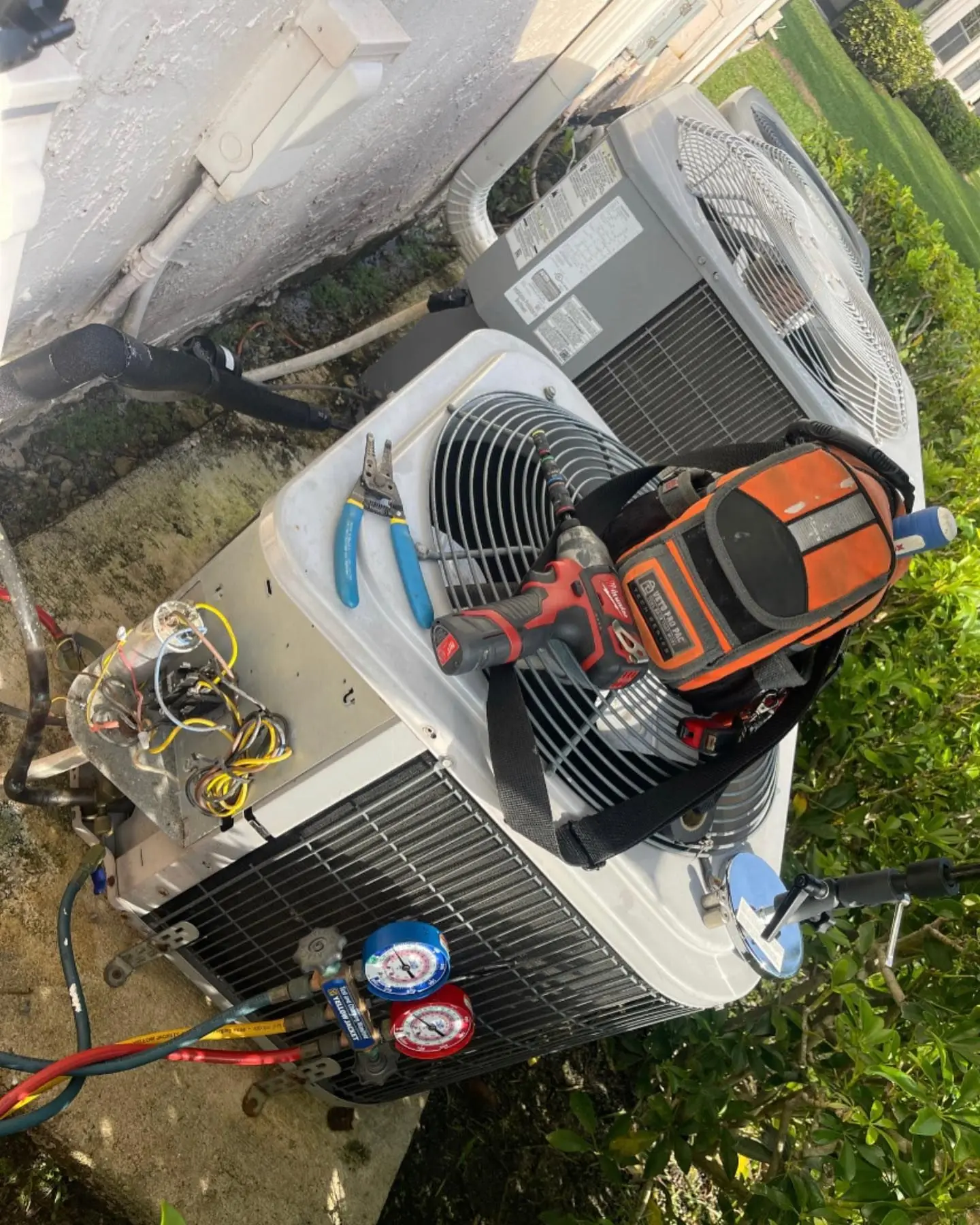 Outdoor condenser opened for service, with refrigerant gauges connected and a tool bag resting on top