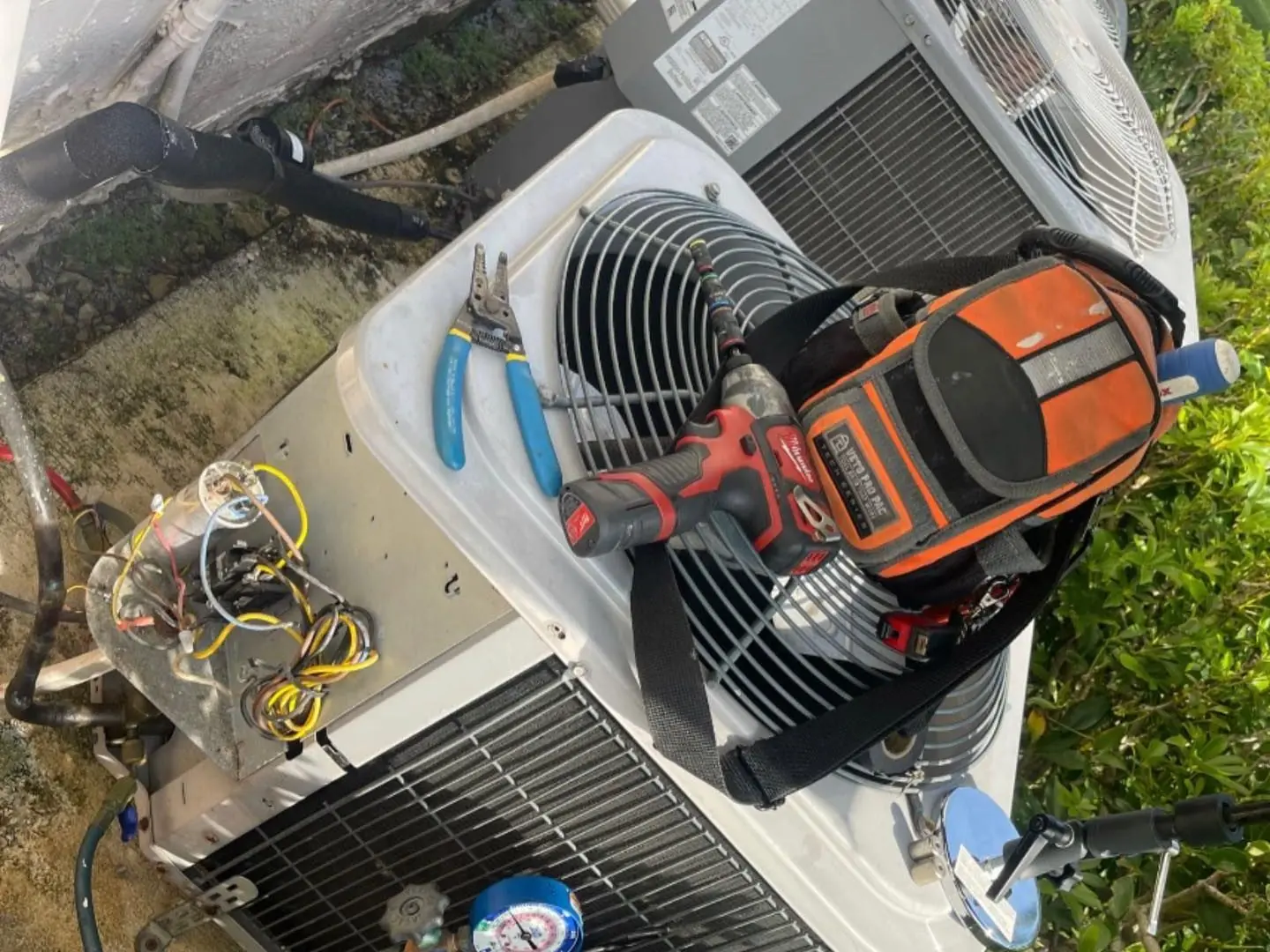 Outdoor condenser opened for service, with refrigerant gauges connected and a tool bag resting on top