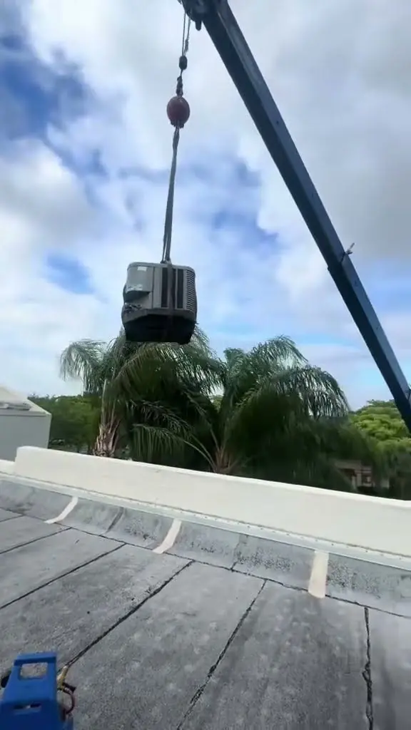 Video thumbnail showing a crane lifting an outdoor AC condensing unit over a flat commercial roof, with palm trees behind