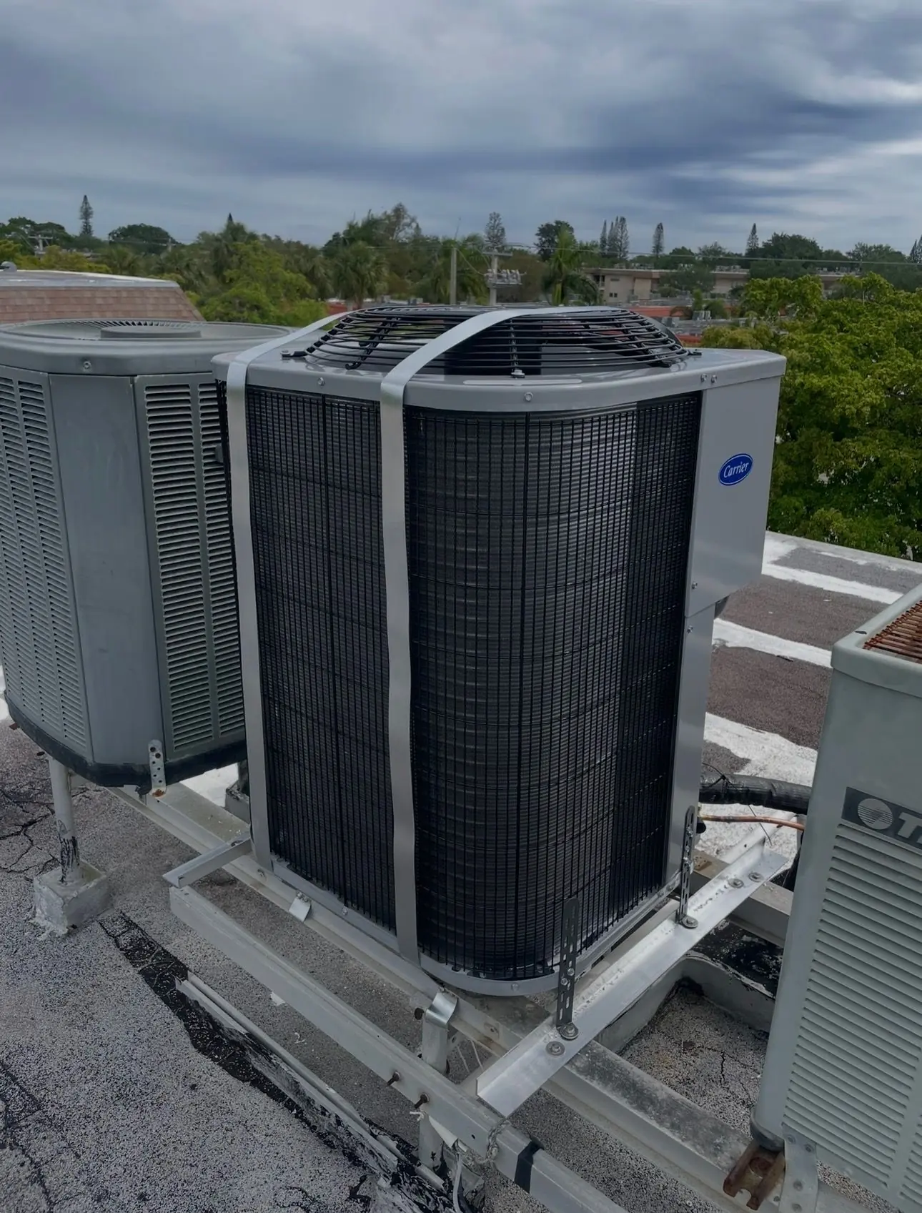 New Carrier condenser on a rooftop equipment stand, held down with metal hurricane straps, beside older units