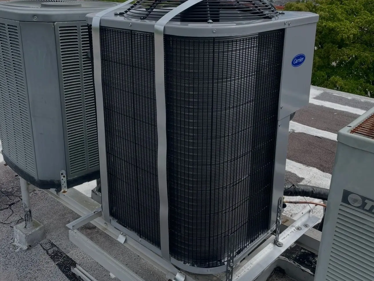New Carrier condenser on a rooftop equipment stand, held down with metal hurricane straps, beside older units