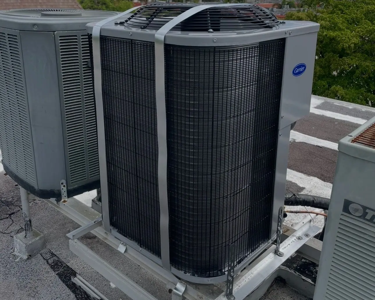 New Carrier condenser on a rooftop equipment stand, held down with metal hurricane straps, beside older units