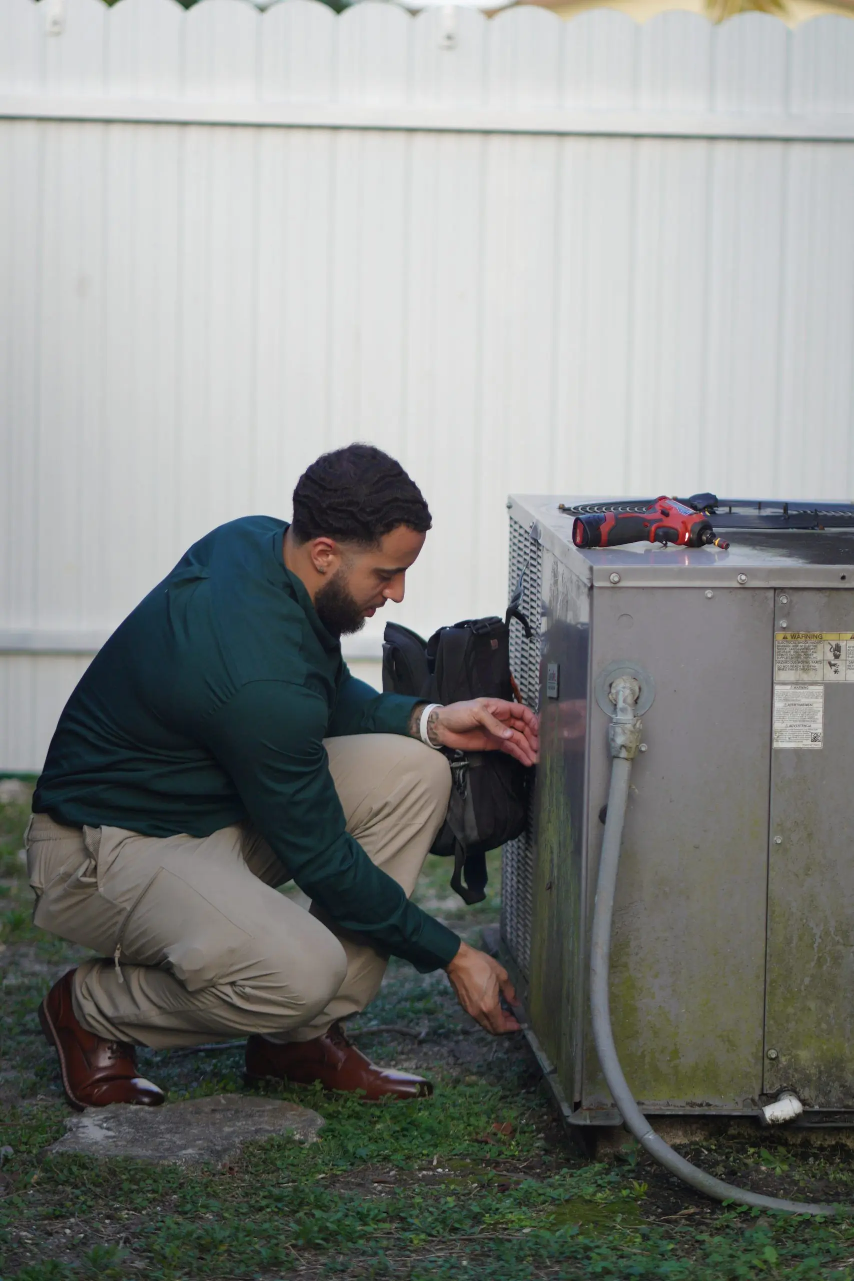 Service Shield technician kneeling at an outdoor unit, reaching into the service panel