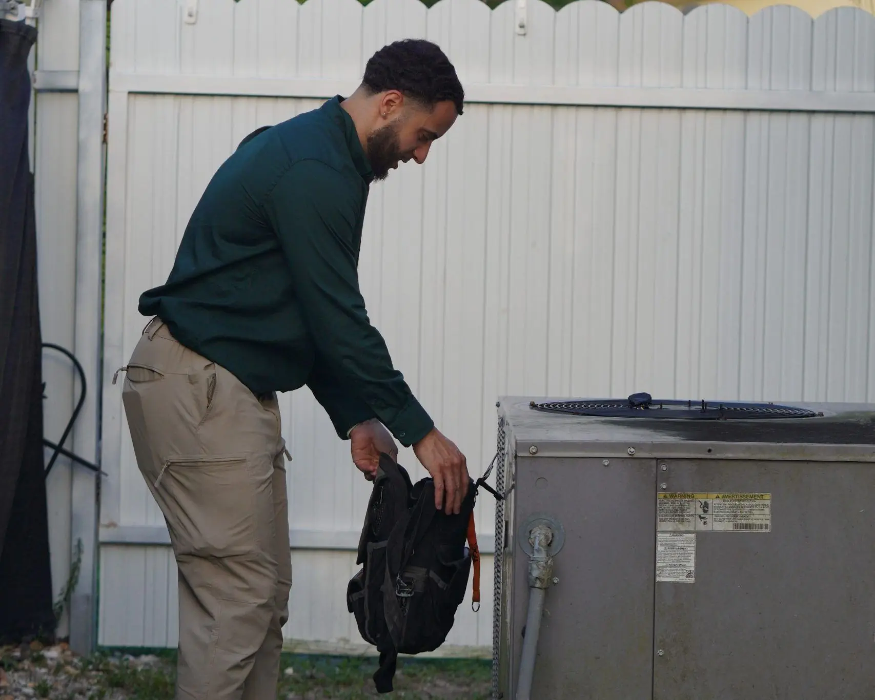 Service Shield technician carrying a tool bag across a backyard toward an outdoor AC condenser beside a white fence