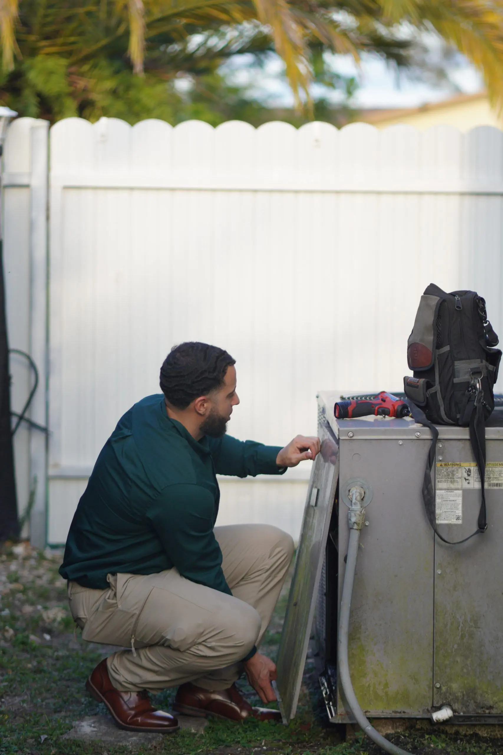 Technician kneeling at an outdoor condenser with its side panel off, checking components inside