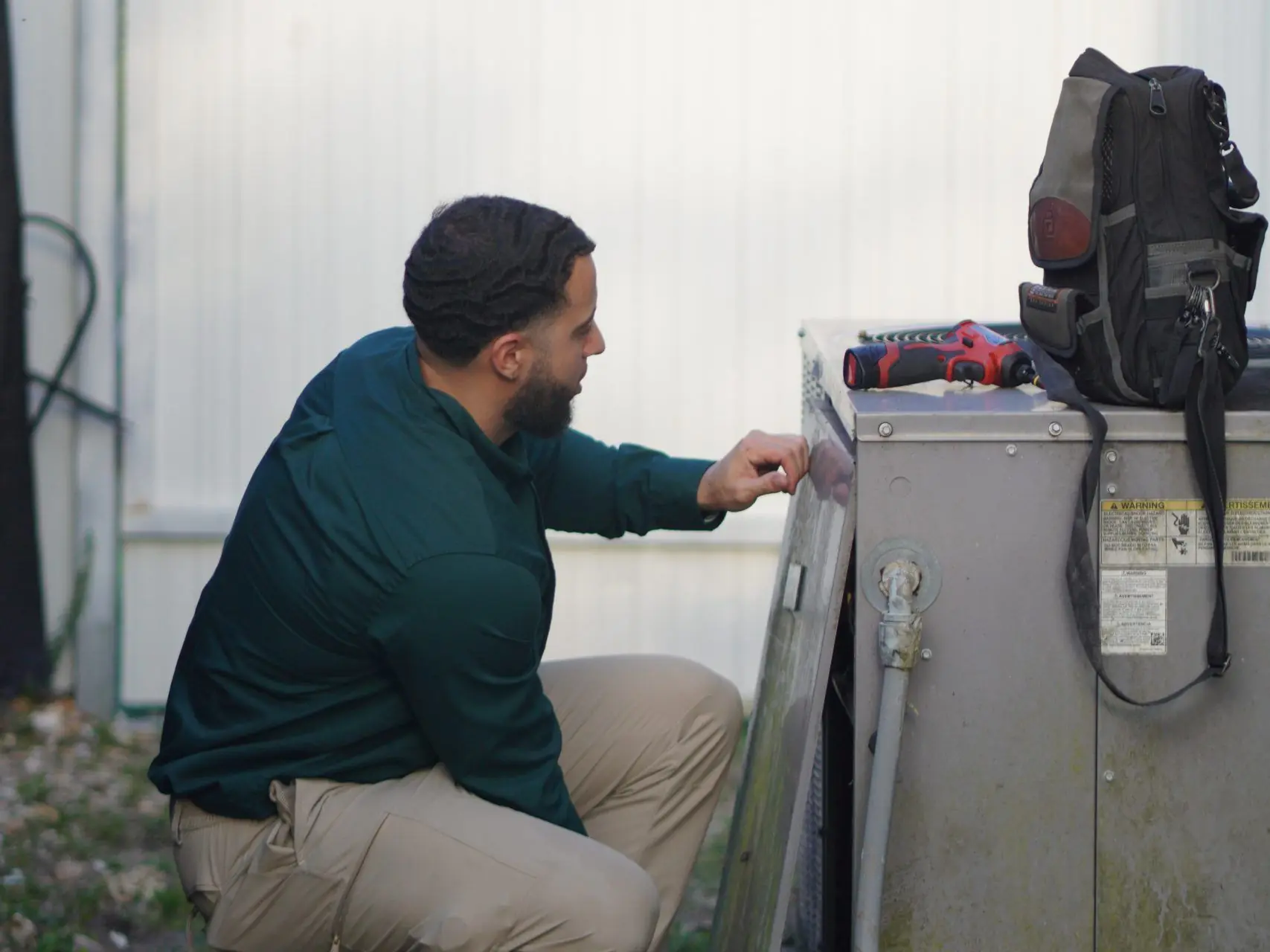 Technician kneeling at an outdoor condenser with its side panel off, checking components inside