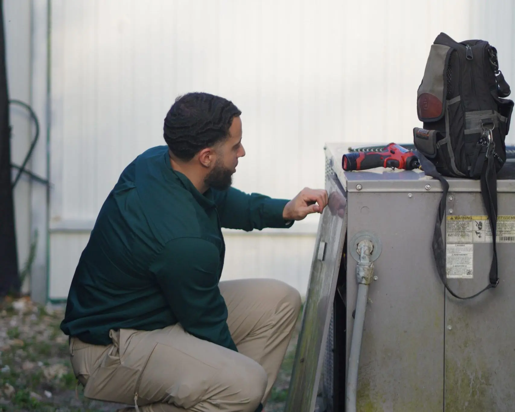 Technician kneeling at an outdoor condenser with its side panel off, checking components inside