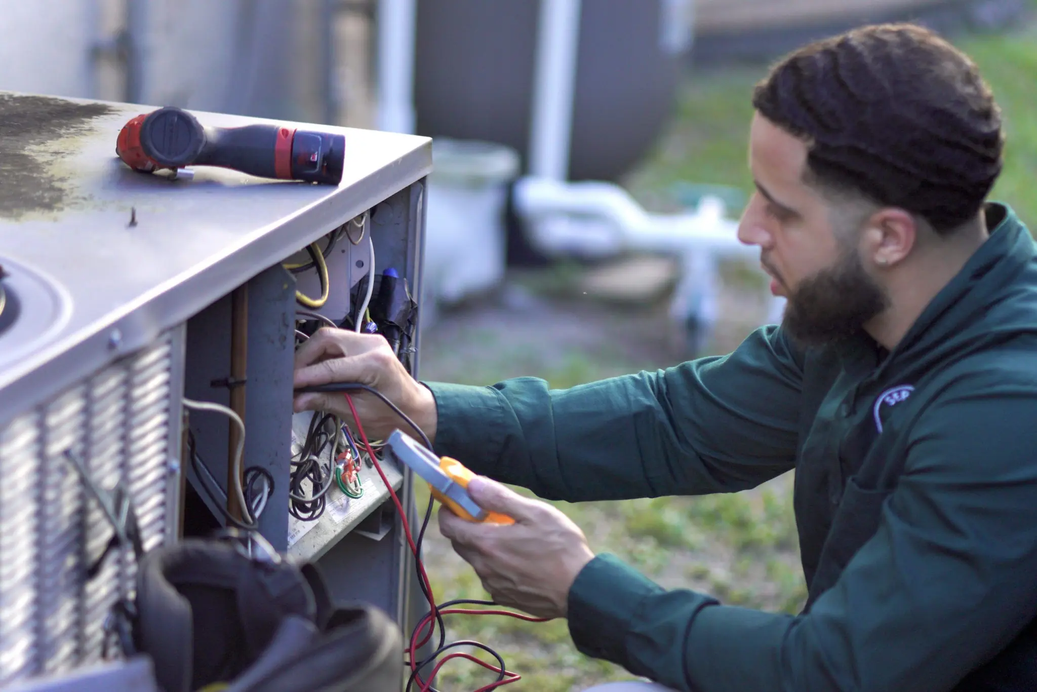 Technician testing wiring inside an outdoor condenser's electrical compartment with a multimeter