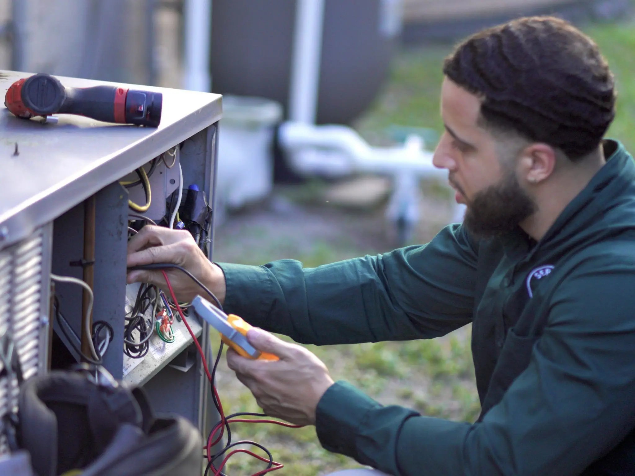 Technician testing wiring inside an outdoor condenser's electrical compartment with a multimeter