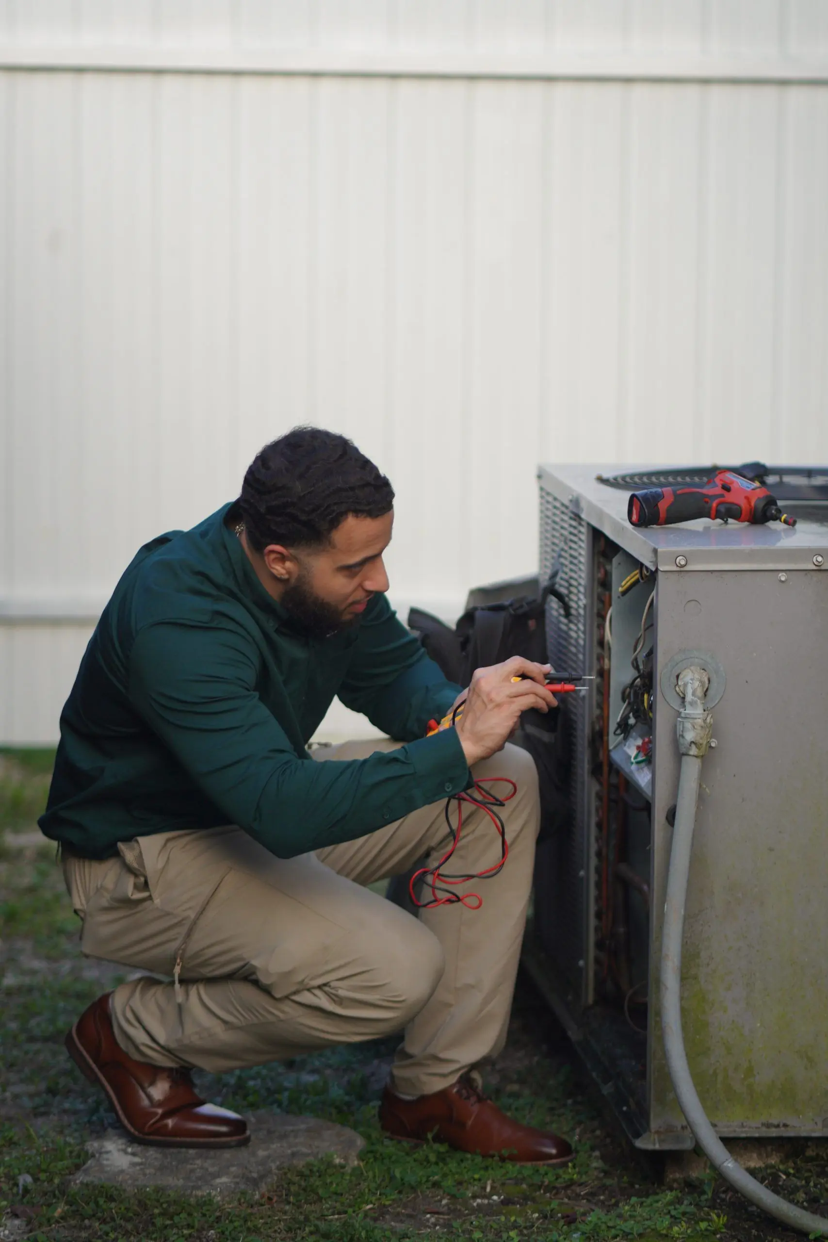 Technician crouched at an open condenser electrical compartment, testing the wiring with meter leads