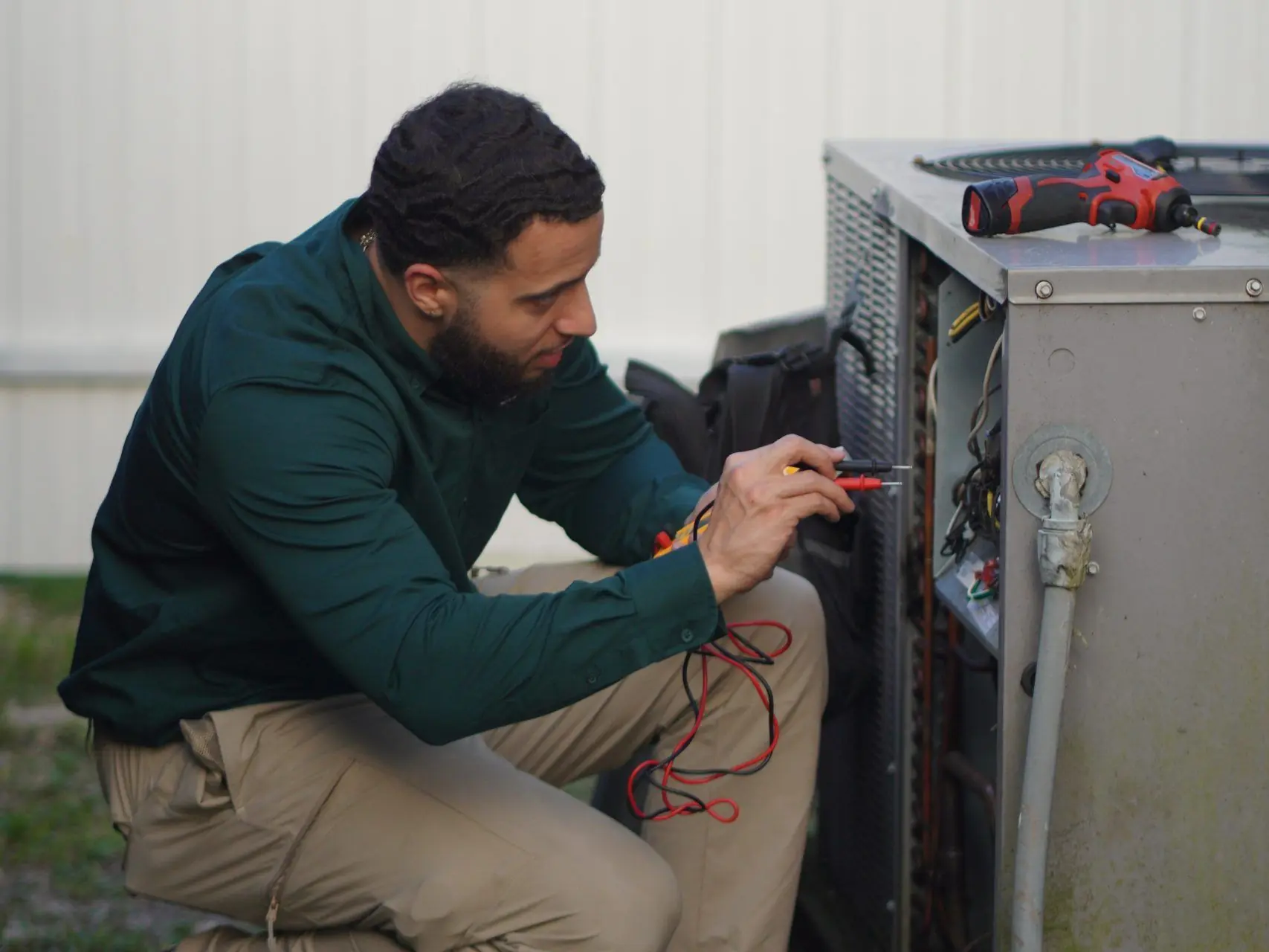 Technician crouched at an open condenser electrical compartment, testing the wiring with meter leads